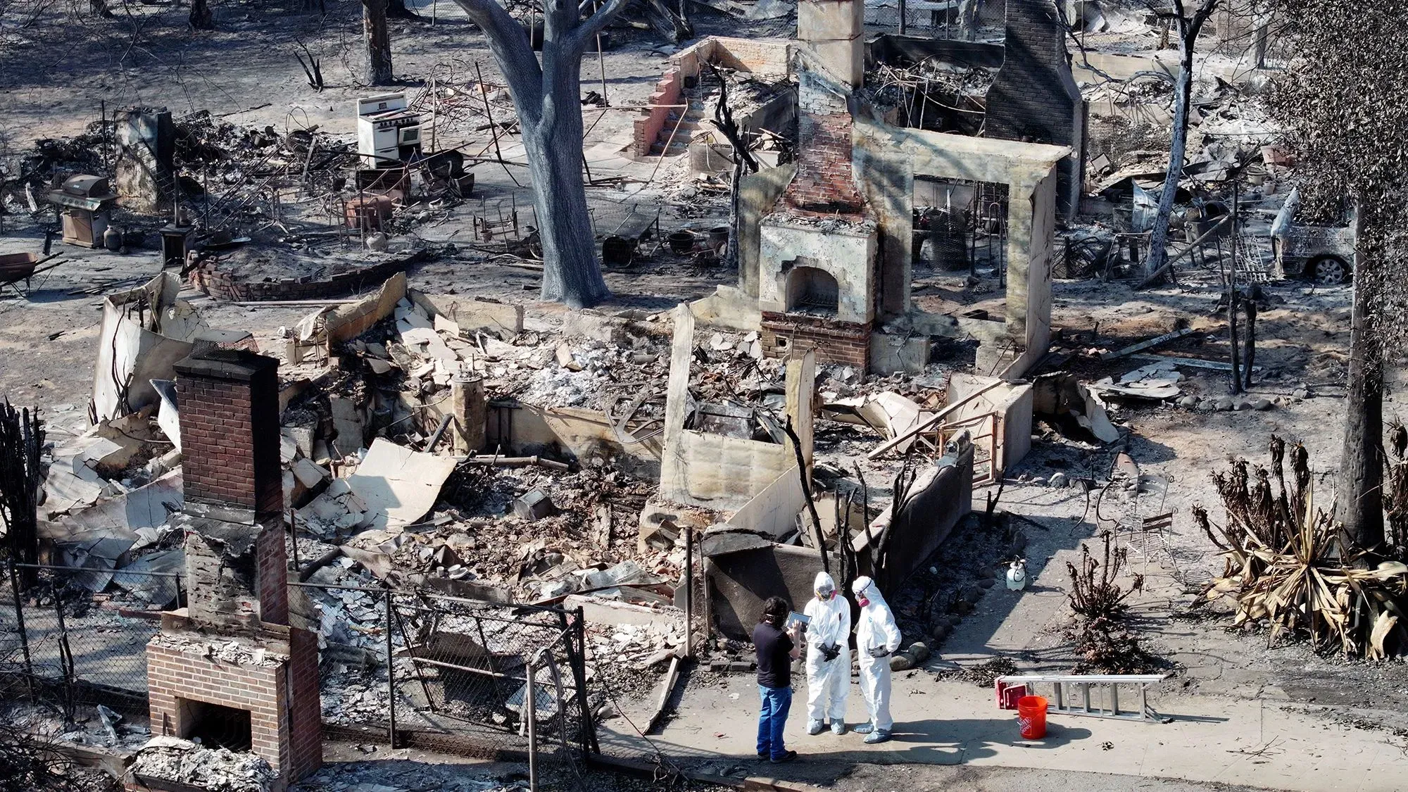 موقع حريق مدمر في كاليفورنيا، يظهر بقايا المنازل المحترقة، مع أشخاص يرتدون ملابس واقية يتحدثون في الموقع.