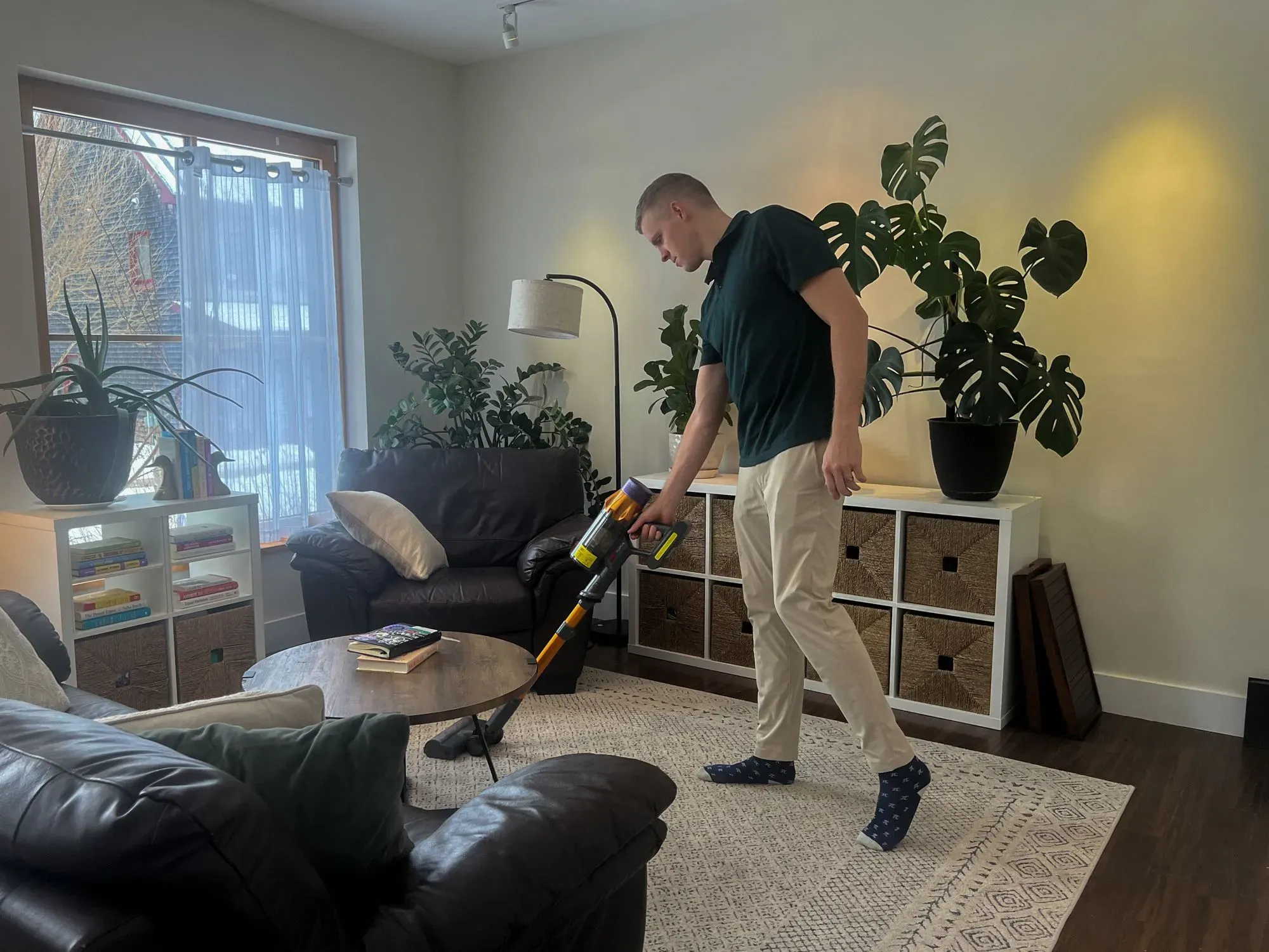 رجل يستخدم مكنسة كهربائية في غرفة معيشة أنيقة، محاط بأثاث مريح ونباتات داخلية، مما يعكس أهمية المشاركة في الأعمال المنزلية.