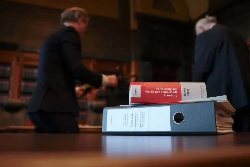 كتب قانونية ومجلدات في قاعة محكمة، مع محامين يتحركون في الخلفية، تعكس أجواء المناقشات القانونية حول حزب البديل من أجل ألمانيا.