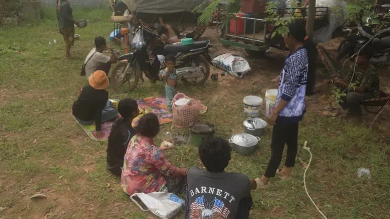 مجموعة من النازحين الكمبوديين يجلسون في منطقة مفتوحة، يتشاركون الطعام ويعبرون عن قلقهم بشأن نقص الإمدادات بعد الاشتباكات على الحدود مع تايلاند.