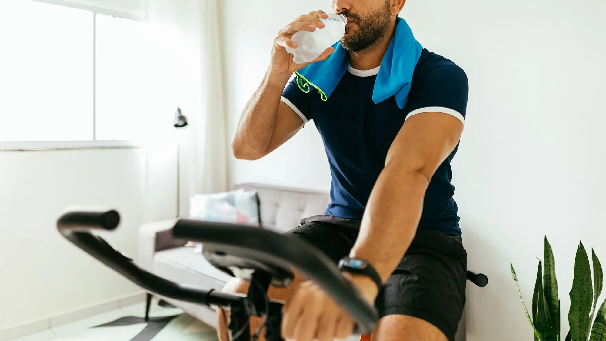 رجل يشرب الماء أثناء ركوبه على دراجة ثابتة في غرفة مضاءة جيدًا، مع مناشف حول عنقه ونباتات في الخلفية.
