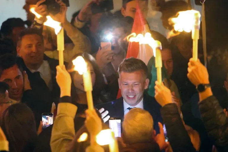 احتفال حاشد بعد فوز بيتر مجار في الانتخابات، حيث يرفع المشاركون الشموع ويهتفون، مع وجود علم المجر في الخلفية.