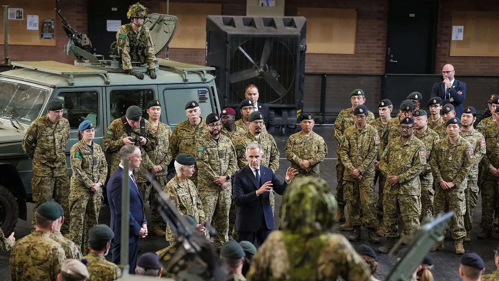 رئيس الوزراء الكندي مارك كارني يتحدث أمام جنود في قاعدة عسكرية، مع مركبة عسكرية في الخلفية، خلال مناقشات حول زيادة الإنفاق الدفاعي.