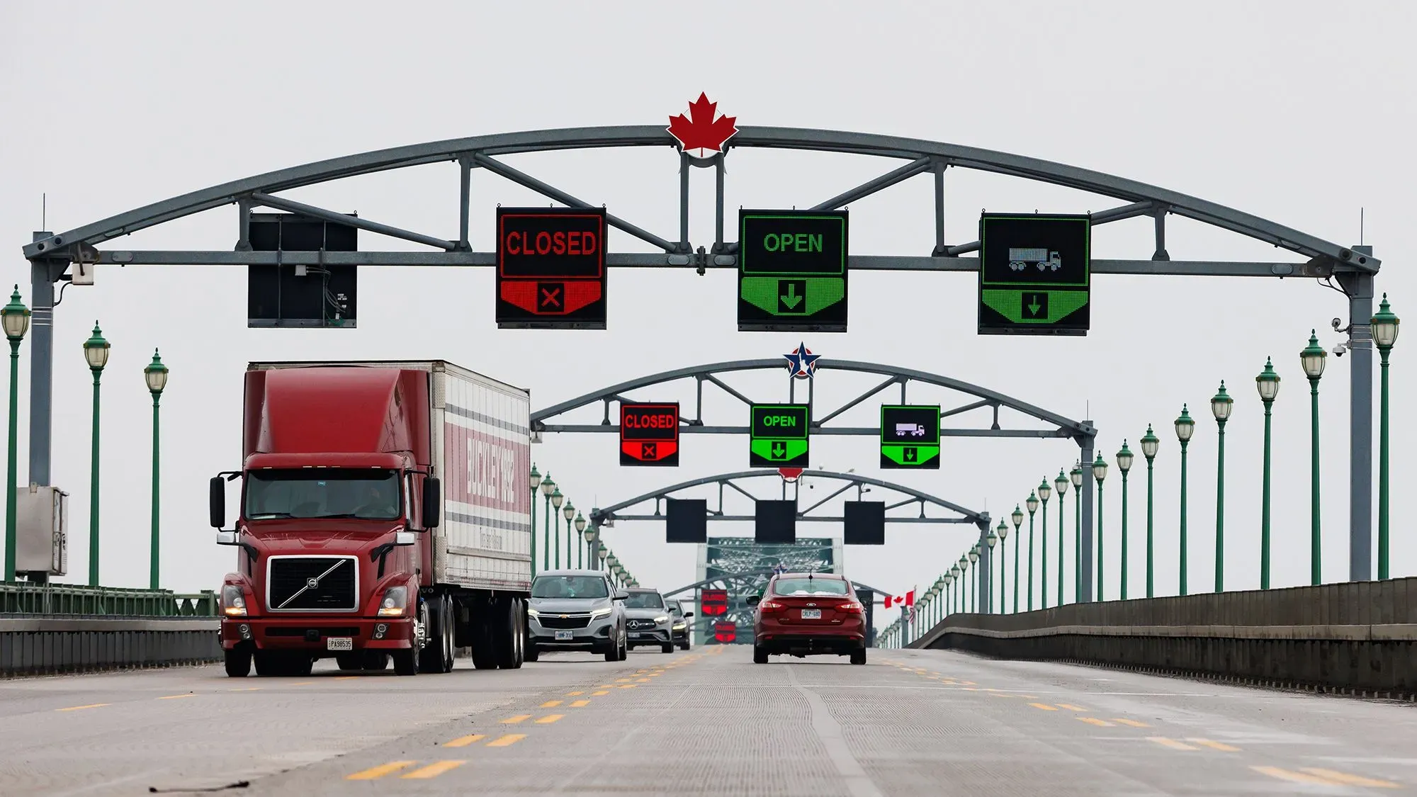 جسر يربط كندا بالولايات المتحدة، يظهر لافتات تشير إلى حالة المرور، مع شاحنة وسيارات تسير في الاتجاهين، في سياق التوترات التجارية.