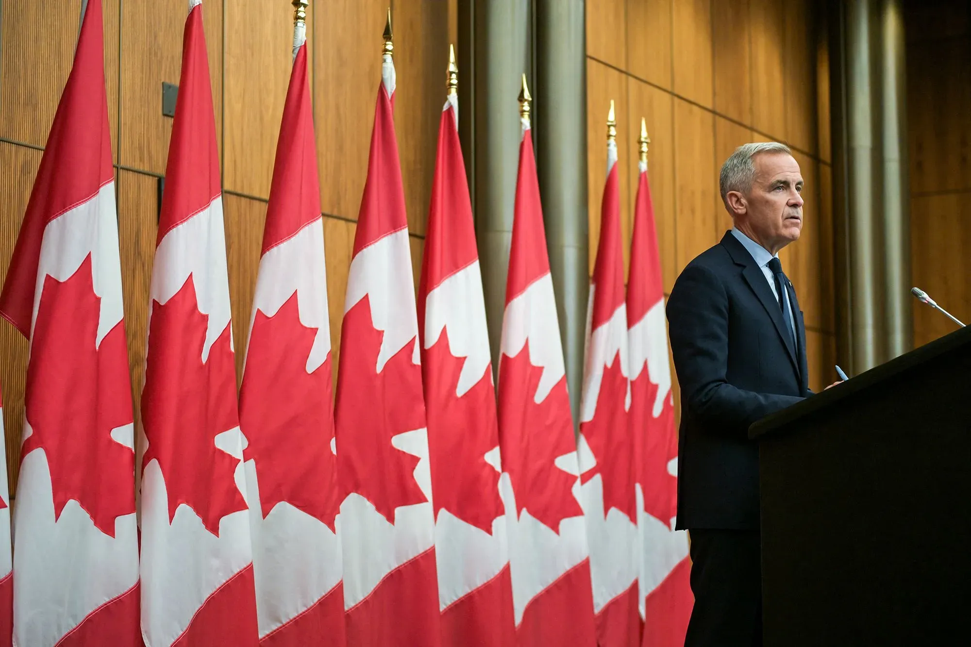 مارك كارني يتحدث أمام مجموعة من الأعلام الكندية، في سياق اجتماعات مع ترامب حول التحديات الاقتصادية والتعريفات الجمركية.