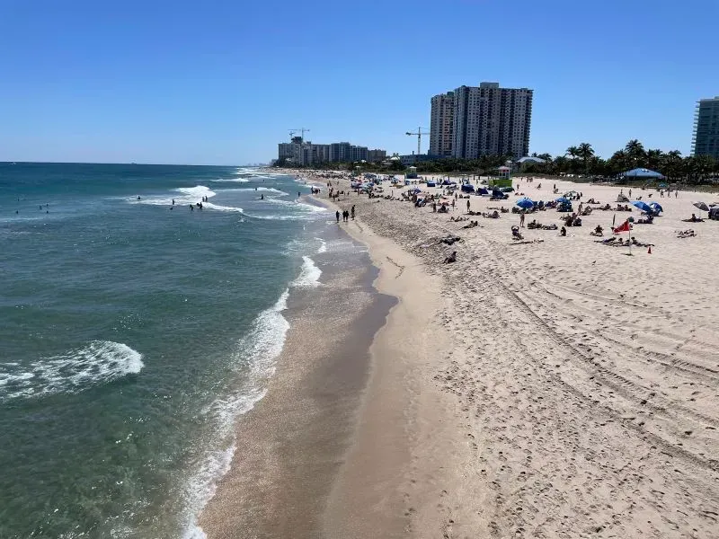 شاطئ مشمس في ميامي مع زوار يستمتعون بالرمال والماء، بينما تظهر المباني الشاهقة في الخلفية، تعكس تزايد اهتمام الكنديين بالعقارات الأمريكية.