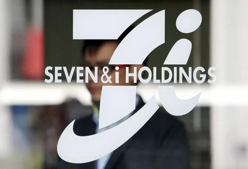 شعار شركة Seven & i Holdings يظهر في واجهة زجاجية، مع شخص يقف في الخلفية، مما يعكس الوضع الحالي في سوق التجزئة.