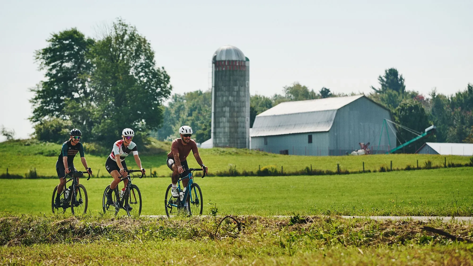 راكبو دراجات ثلاث يتنقلون عبر مزارع الكروم في منطقة البلدات الشرقية بكندا، مع مناظر طبيعية خضراء ومباني زراعية في الخلفية.