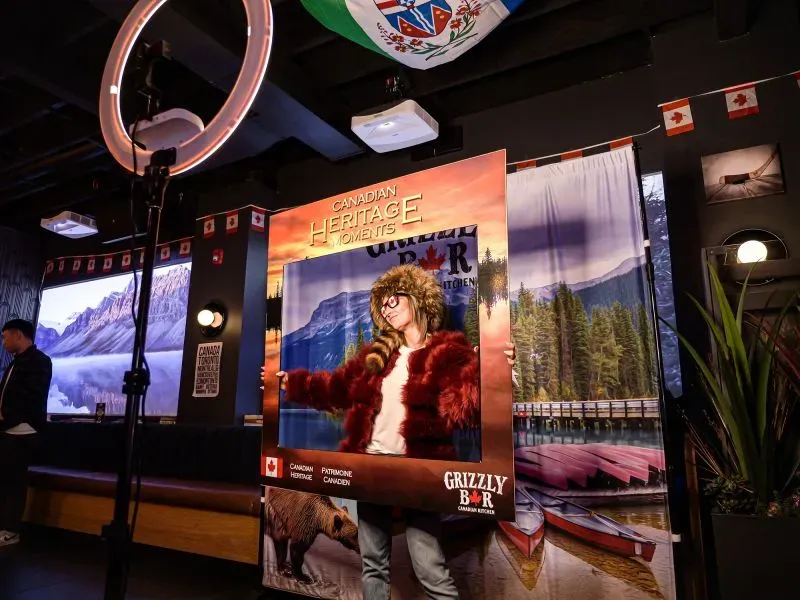 امرأة ترتدي زيًا كنديًا تقف داخل إطار يحمل شعار "لحظات التراث الكندي" في مطعم، مع صور طبيعية خلفها، تعكس الثقافة الكندية.