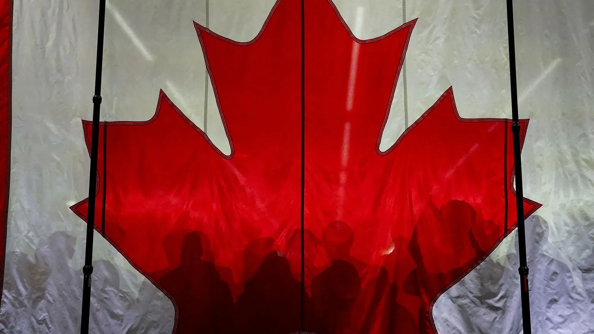 علم كندا الكبير باللونين الأحمر والأبيض، مع ظلال لأشخاص يقفون خلفه، يرمز إلى الانتخابات الكندية وتأثيرها السياسي.