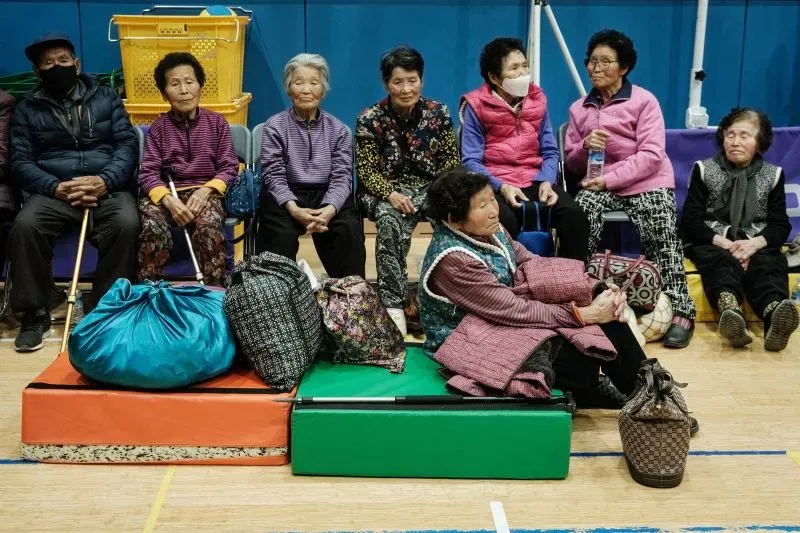 مجموعة من كبار السن يجلسون في مركز إيواء بعد إخلائهم بسبب حرائق الغابات في كوريا الجنوبية، مع حقائب وأغراض شخصية بجانبهم.