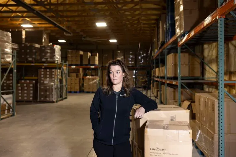 امرأة تقف في مستودع لشركة Busy Baby، محاطة بصناديق البضائع. تعكس الصورة التحديات التي تواجهها الشركات الصغيرة في ظل الرسوم الجمركية المتزايدة.