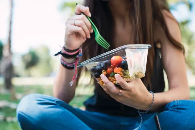 امرأة تأكل سلطة تحتوي على طماطم وفواكه في عبوة بلاستيكية، في حديقة، تعكس أهمية التغذية وتأثير المواد الكيميائية على الصحة.