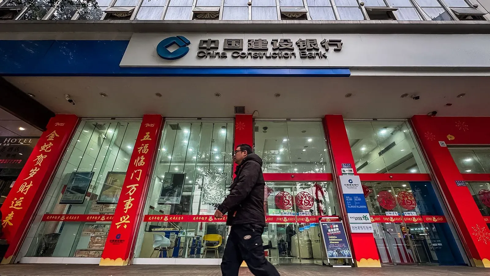 واجهة بنك الصين الإنشائي مع لافتات حمراء، حيث يمر شخص بجانب المدخل، في سياق جمع البنوك المملوكة للدولة لرأس المال لتعزيز الاقتصاد.