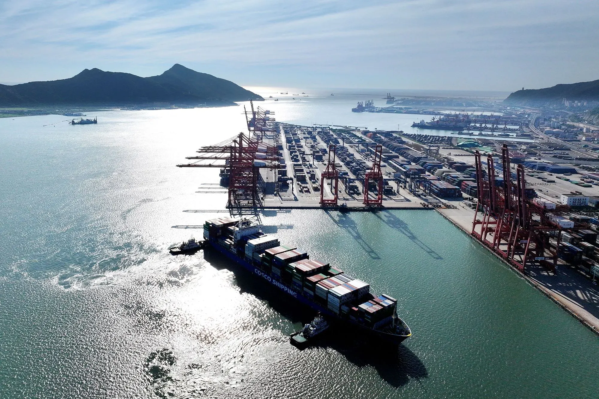 صورة جوية لميناء مزدحم، تُظهر سفينة شحن محملة بالحاويات، مع رافعات ومرافق تخزين في الخلفية، تعكس أهمية التجارة بين الصين والولايات المتحدة.