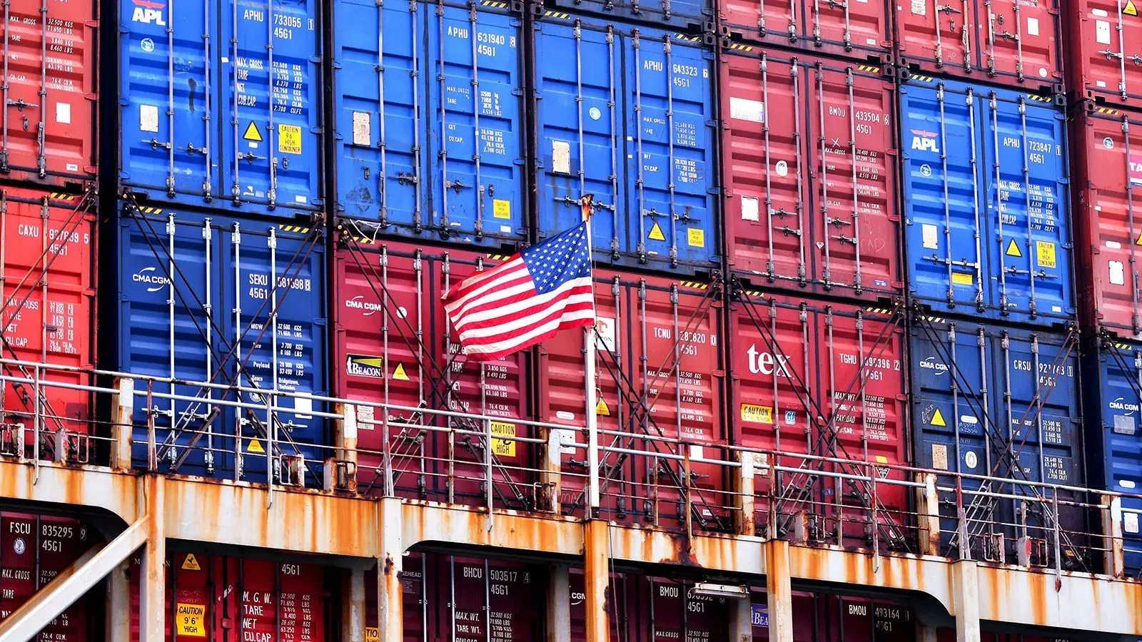 أعلام الولايات المتحدة تتدلى بين حاويات شحن ملونة، في سياق التوترات التجارية بين الصين والولايات المتحدة.