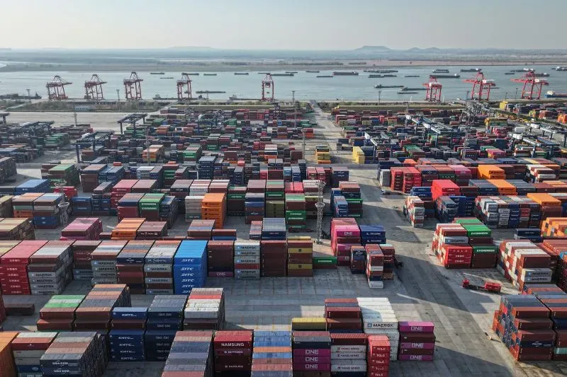صورة لميناء حاويات مزدحم يحتوي على العديد من الحاويات الملونة، مع رافعات في الخلفية، تعكس تأثير الحرب التجارية بين الولايات المتحدة والصين.