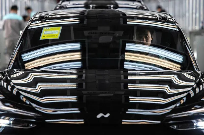 شخص يجلس داخل سيارة كهربائية سوداء في مصنع، مع انعكاسات الأضواء الساطعة على الزجاج، مما يعكس تقدم الصين في صناعة السيارات الكهربائية.