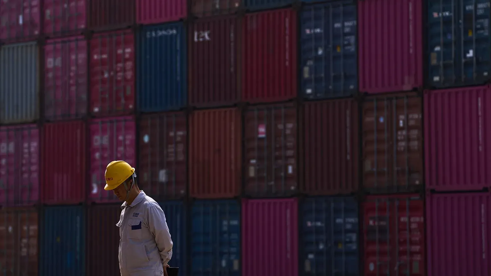 عامل يرتدي خوذة صفراء يقف أمام حاويات شحن ملونة في ميناء، مما يبرز التوترات التجارية بين الصين والولايات المتحدة.