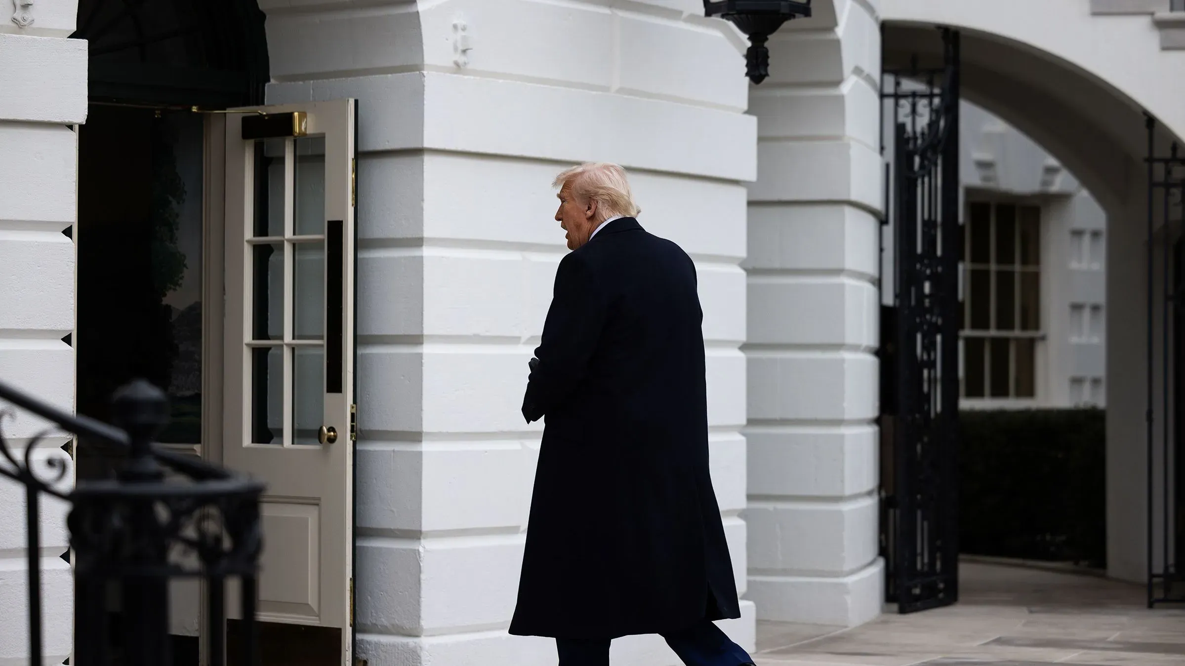 رجل يرتدي معطفًا طويلًا يغادر البيت الأبيض، مع التركيز على الباب المفتوح خلفه، مما يعكس التغيرات في السياسة الخارجية الأمريكية.