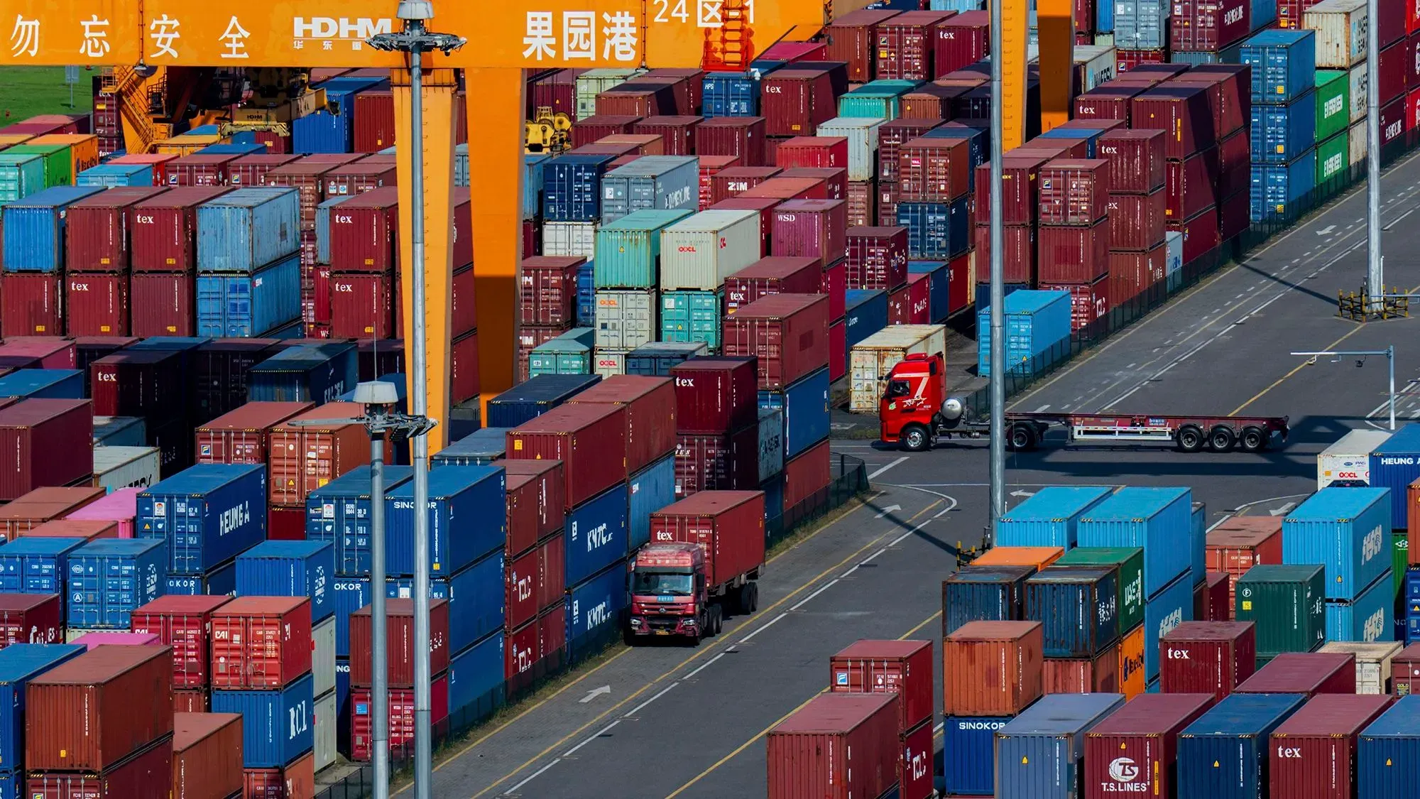 صورة لميناء بحري مزدحم، حيث تتراص حاويات الشحن الملونة بشكل كثيف، مع شاحنة تسير بين الحاويات. تعكس الصورة حجم التجارة العالمية والتحديات الاقتصادية.