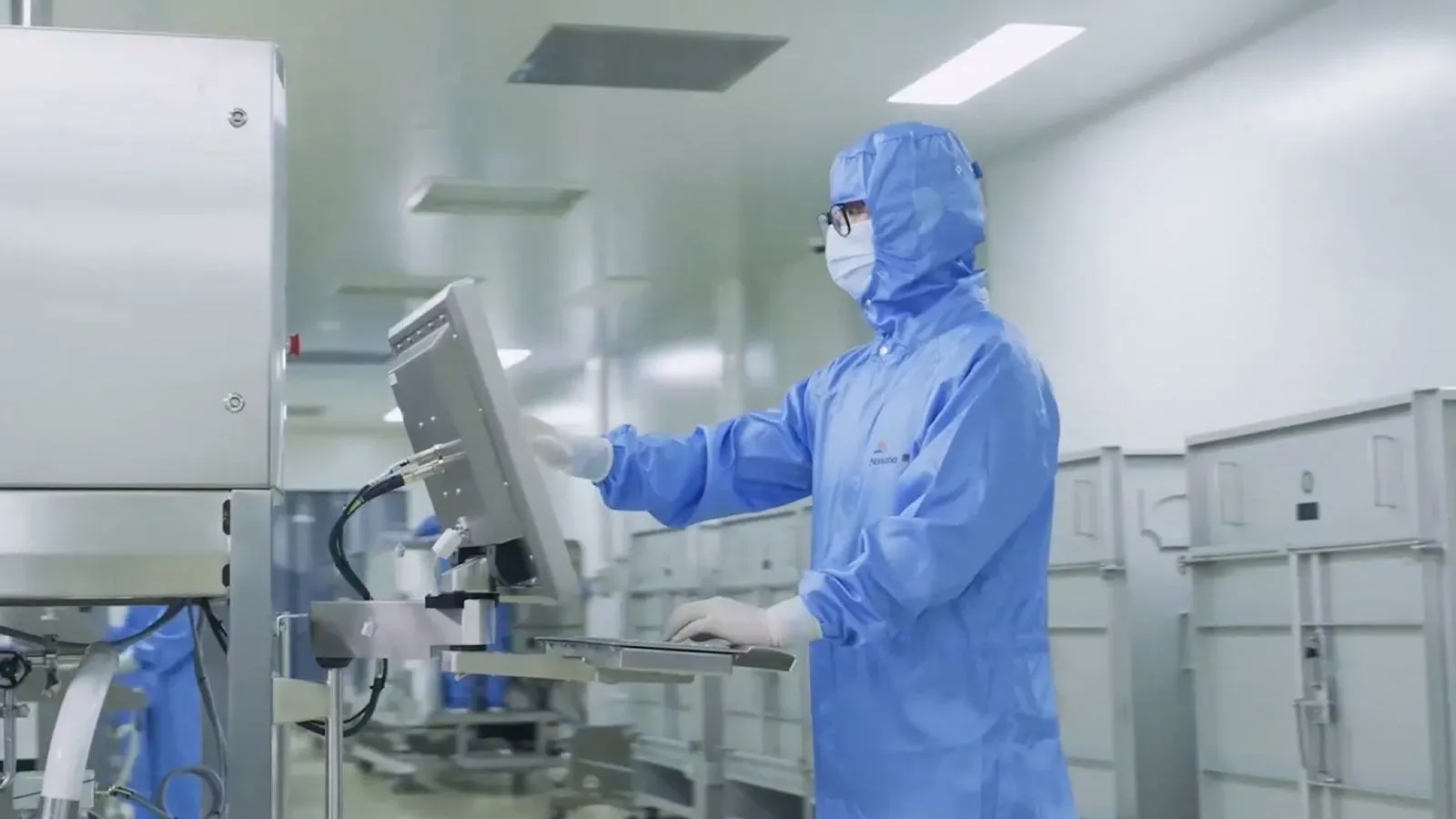 عامل يرتدي بدلة واقية زرقاء في مختبر حديث، يتفاعل مع جهاز كمبيوتر، مما يعكس الابتكارات في صناعة الأدوية الصينية.