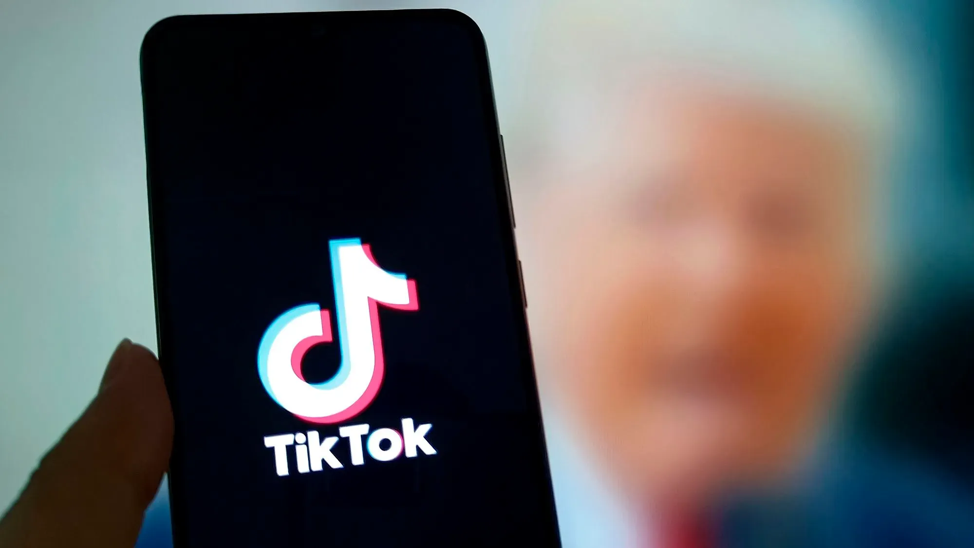 شاشة هاتف تعرض شعار تطبيق تيك توك، مع صورة غير واضحة خلفية لرجل يتحدث، تعكس الأجواء المحيطة بمفاوضات التطبيق.