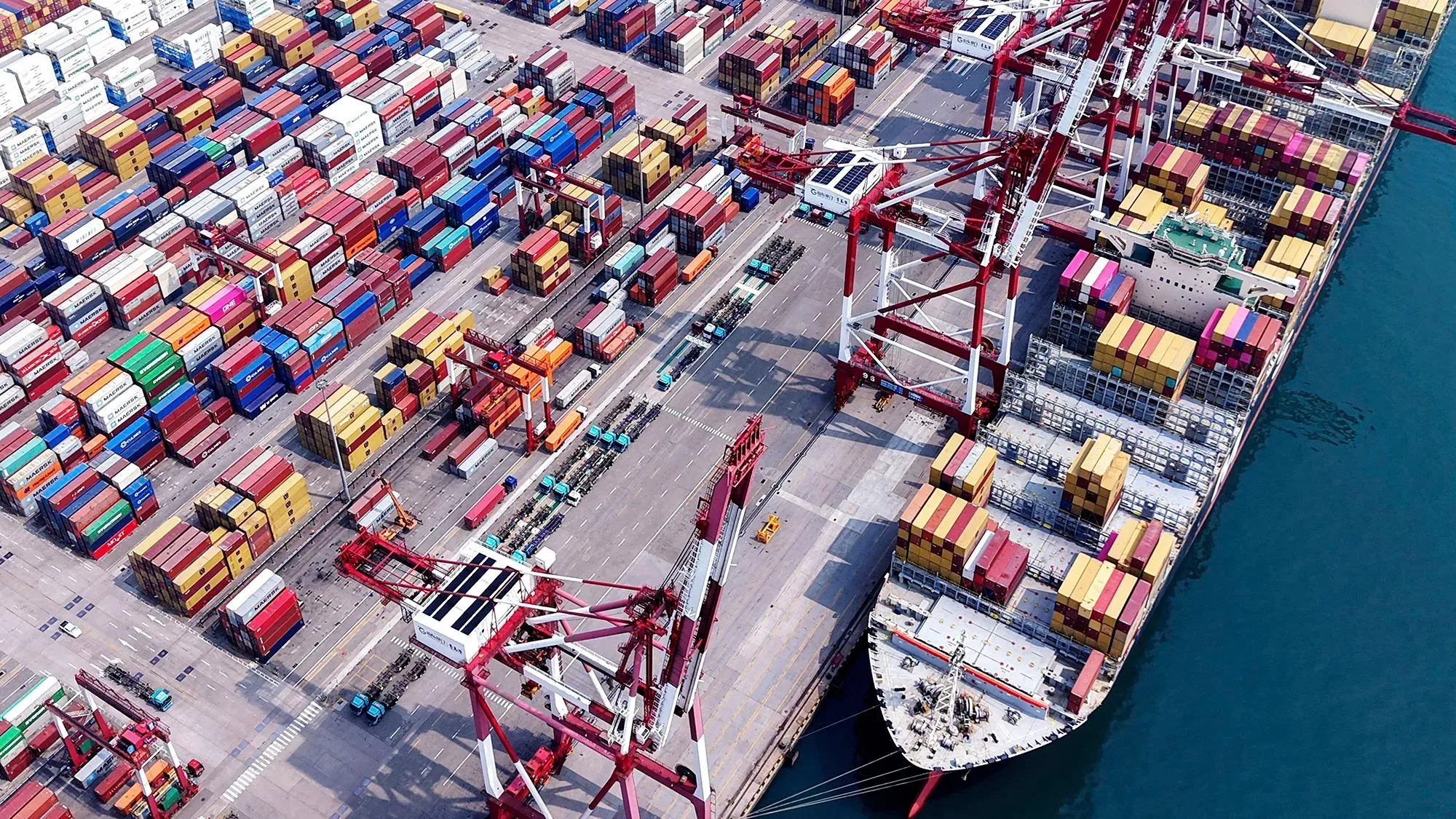 صورة جوية لميناء بحري مزدحم يحتوي على حاويات ملونة وسفينة شحن كبيرة، تعكس تأثير التعريفات الجمركية على التجارة العالمية.