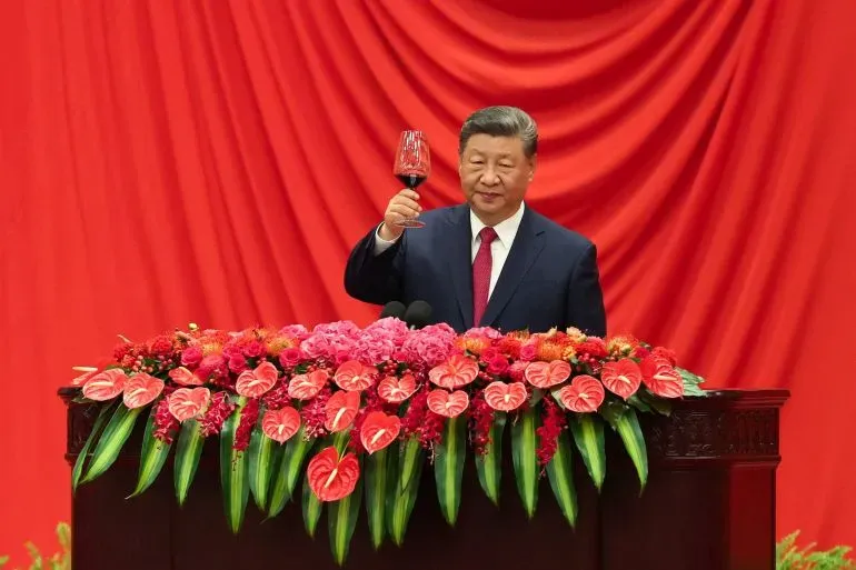 شي جين بينغ يرفع كأسًا خلال حدث رسمي، محاطًا بزهور ملونة وخلفية حمراء، في سياق توترات تجارية مع الولايات المتحدة.