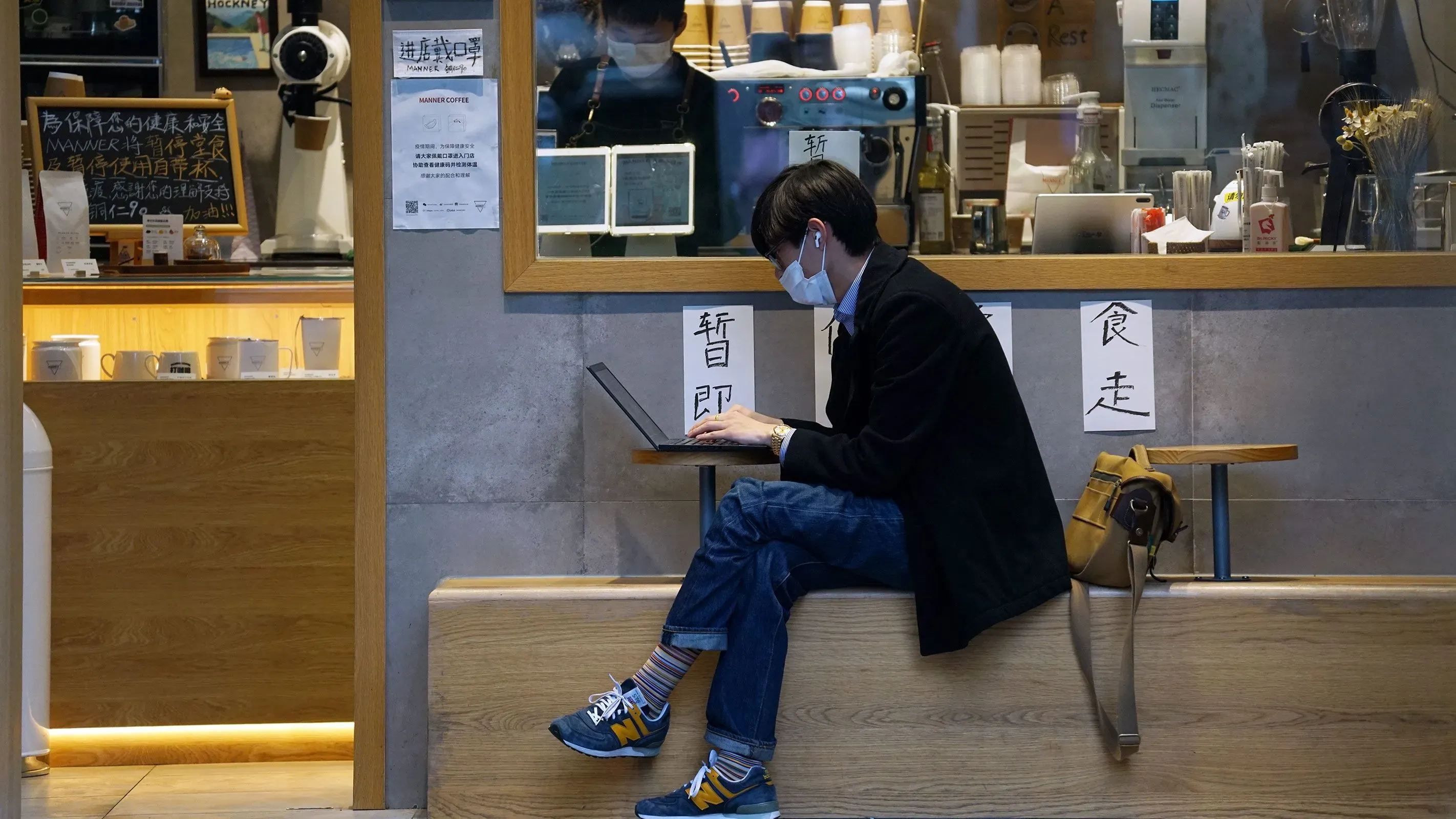 شخص يجلس في مقهى، يرتدي كمامة ويستخدم حاسوبًا محمولًا، مع خلفية تحتوي على لافتات وأدوات قهوة، مما يعكس بيئة العمل الحديثة في الصين.