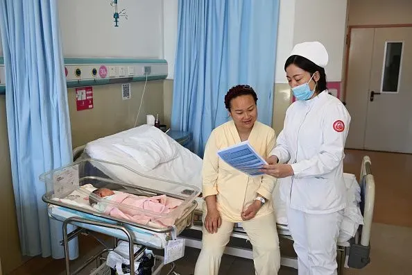 امرأة جالسة بجانب سرير مستشفى، تتحدث مع مربية ترتدي زيًا طبيًا، بينما ينام طفل رضيع في سرير مجاور.