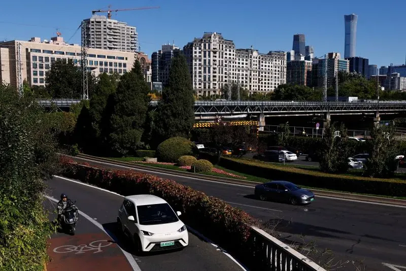 مشهد حضري في بكين يظهر سيارات كهربائية تسير على الطرق بين المباني الحديثة والأشجار، مما يعكس تقدم الصين في صناعة السيارات الكهربائية.