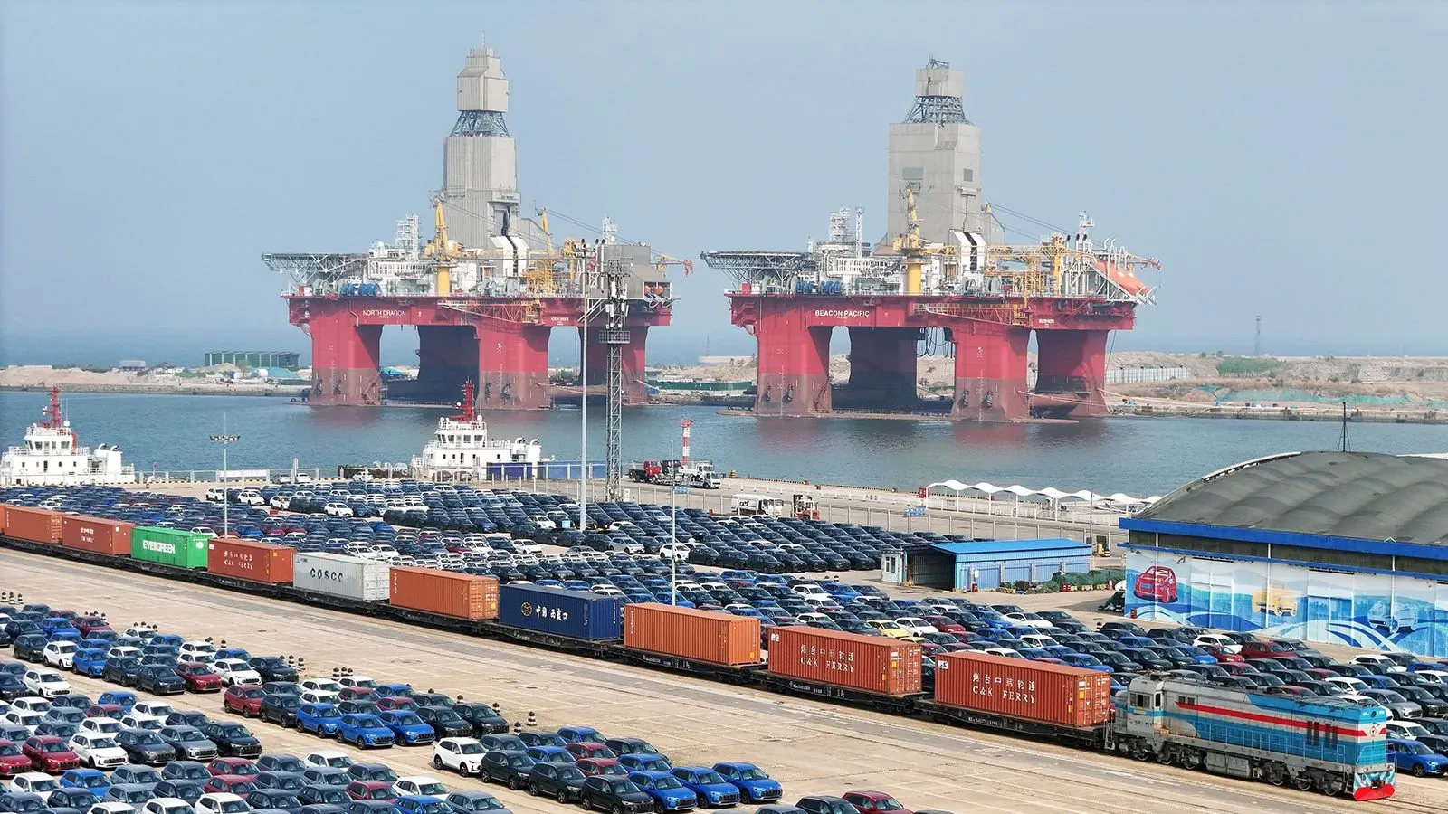 صورة لميناء مزدحم يظهر فيه سفن ضخمة للتنقيب عن النفط، مع شاحنات وقطارات تحمل حاويات، ومركبات متوقفة في الخلفية.
