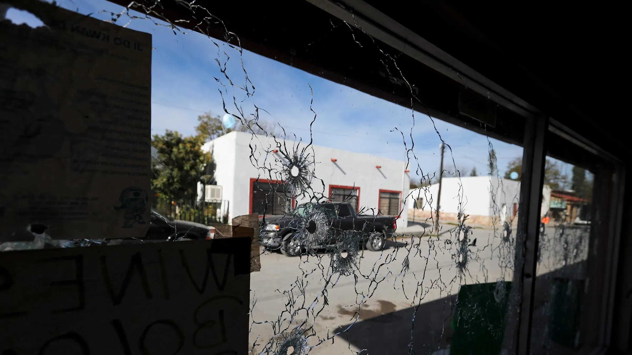 زجاج مكسور يحمل آثار طلقات نارية، يظهر من خلاله مشهد لشارع في المكسيك، مما يعكس تأثير العنف المرتبط بعصابات المخدرات.