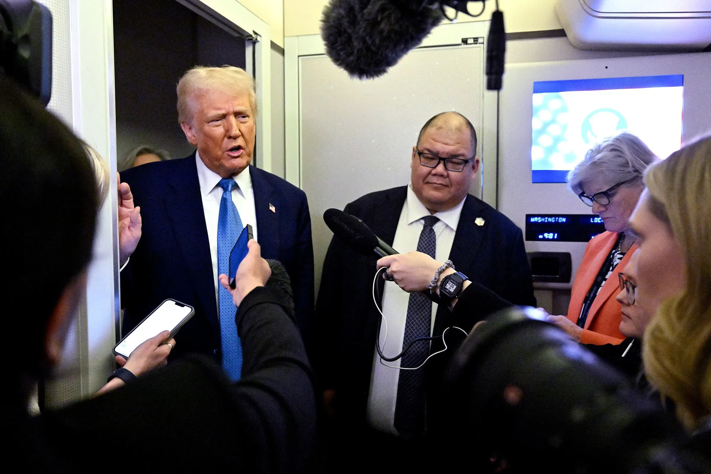 ترامب يتحدث مع الصحفيين في ممر الطائرة، بينما يحيط به عدد من المراسلين، مع وجود شاشات عرض خلفهم.
