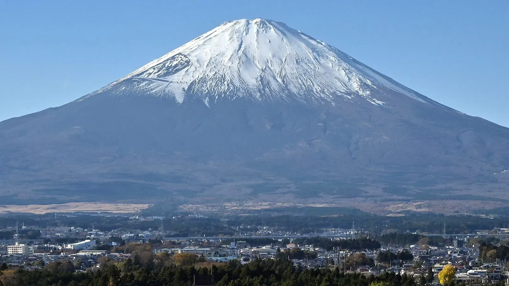 جبل فوجي المغطى بالثلوج، يظهر بوضوح في السماء الزرقاء، محاطًا بالمدن والريف الياباني، مما يعكس جمال الطبيعة والتحديات في التسلق.