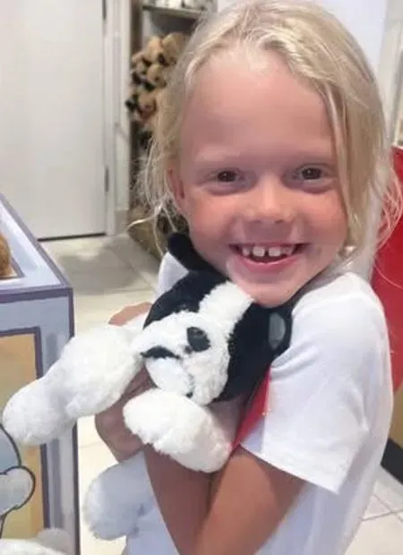 طفلة صغيرة تحمل دمية، مبتسمة وتظهر أسنانها الأمامية المفقودة، تعكس براءة الطفولة وسط أحداث مأساوية في تكساس.