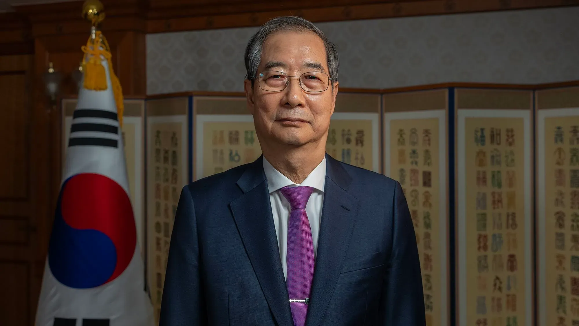 رئيس وزراء كوريا الجنوبية بالوكالة هان داك سو، يقف أمام العلم الكوري الجنوبي وخلفية تقليدية، معبراً عن موقفه من الرسوم الجمركية الأمريكية.
