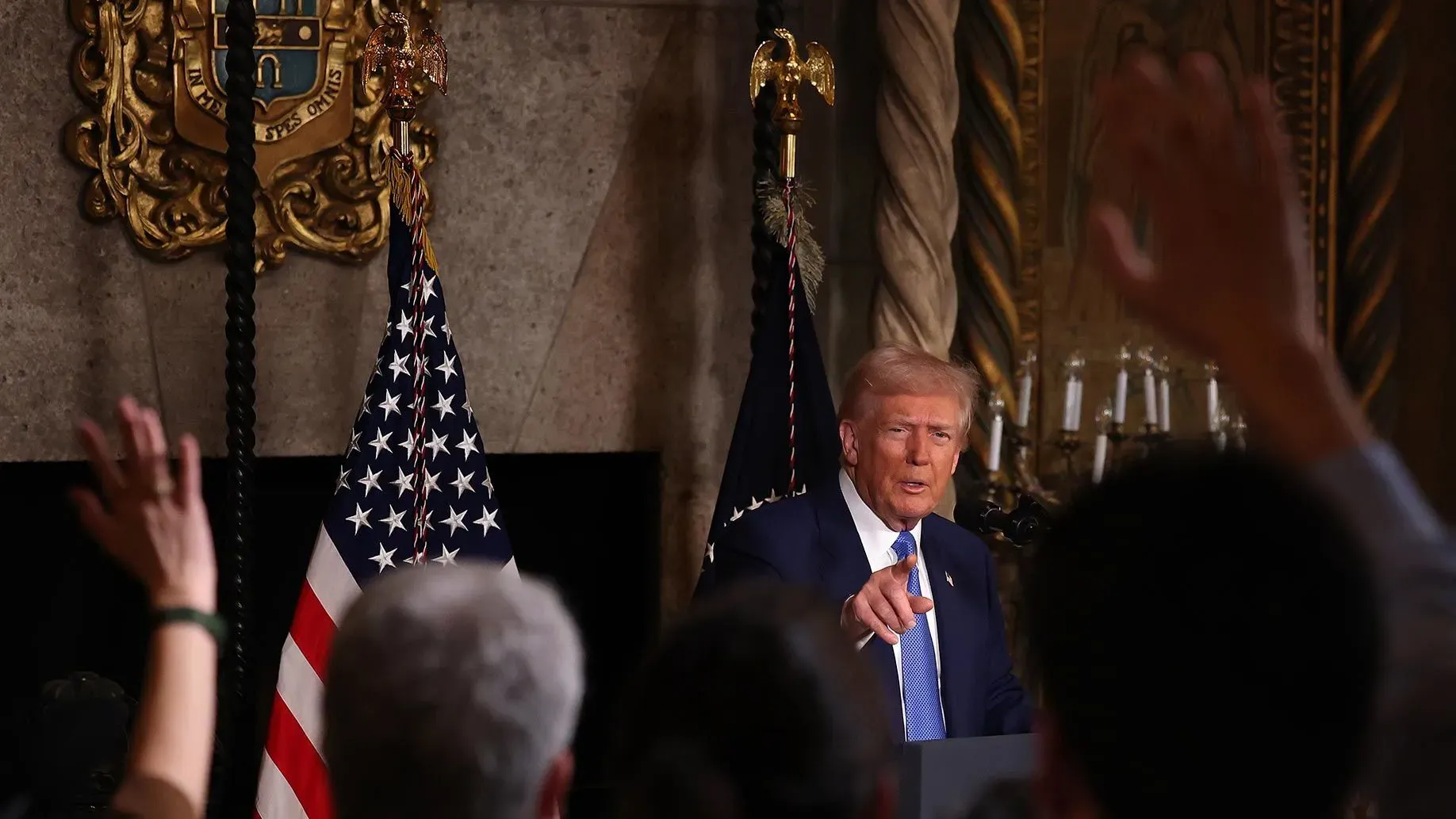 ترامب يتحدث في مؤتمر صحفي، مع رفع الأيدي من الحضور، وعلم الولايات المتحدة خلفه، يعكس تفاعلاته مع الجمهور حول قضايا سياسية.