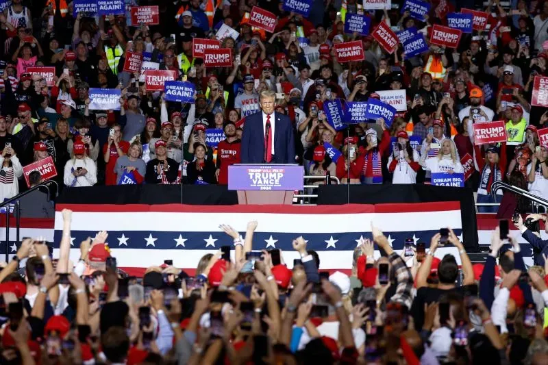 دونالد ترامب يتحدث أمام حشد كبير من أنصاره في تجمع انتخابي، حيث يحمل الجمهور لافتات داعمة ويحضر بأزياء تحمل صورته.