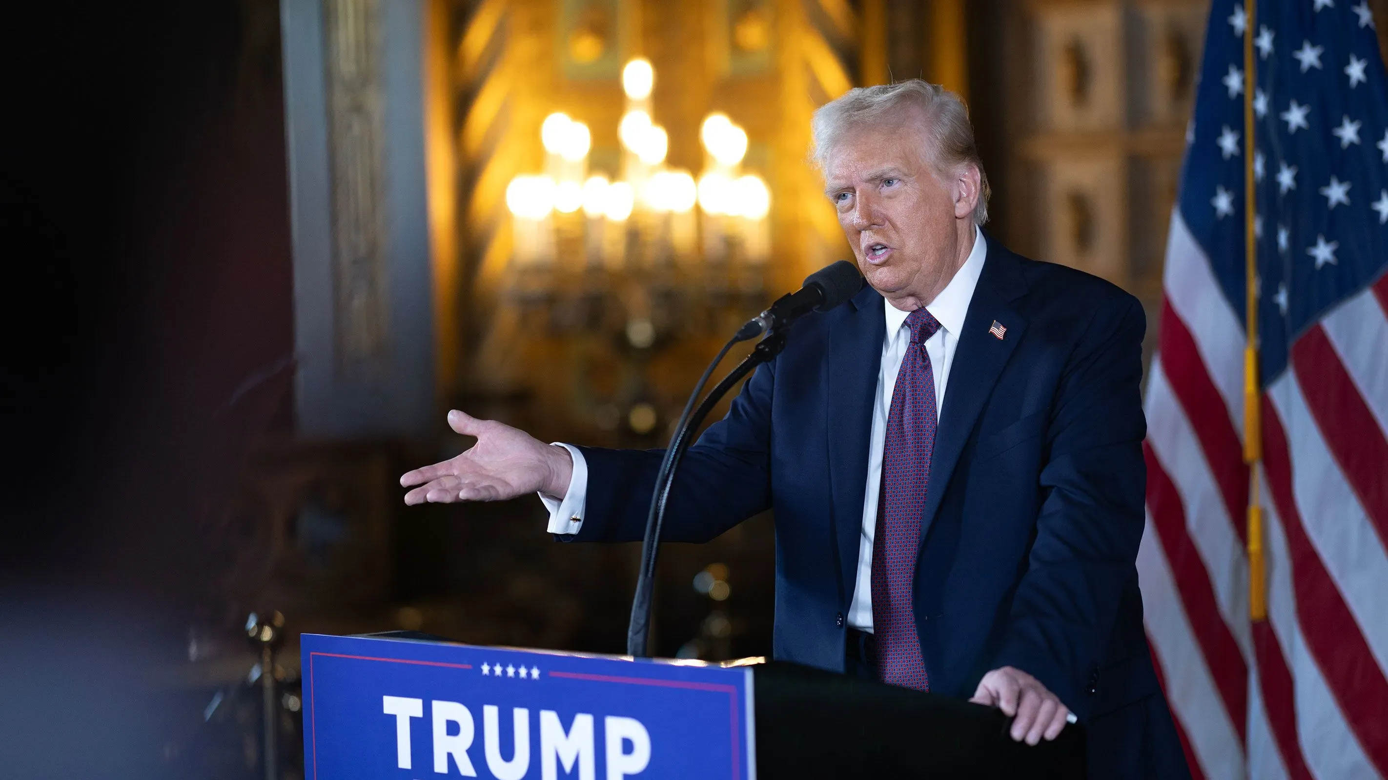 دونالد ترامب يتحدث في مؤتمر صحفي، مع لافتة تحمل اسمه خلفه، وعلم أمريكي يظهر إلى جانبه، في سياق استعداداته لفترة ولايته الثانية.