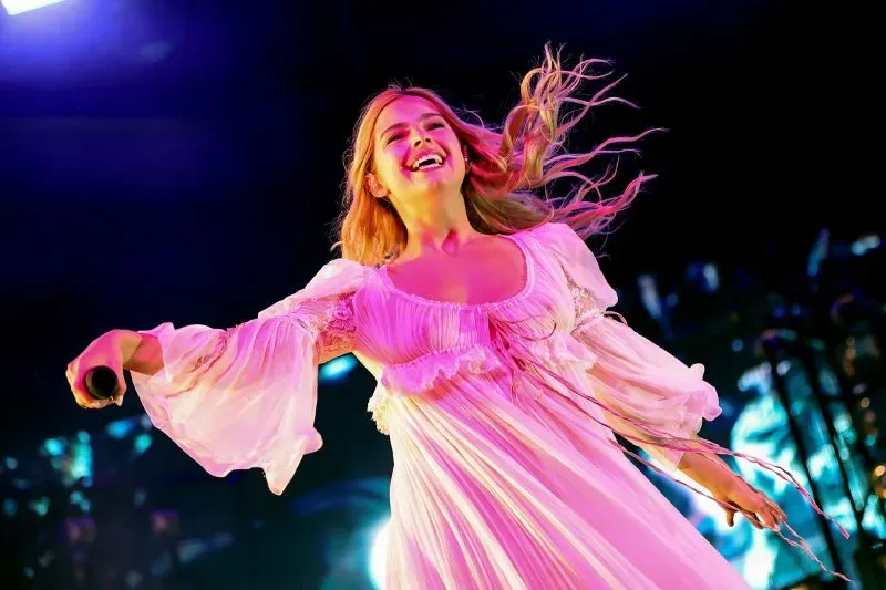 مغنية ترتدي فستانًا أبيض فضفاض وتبتسم أثناء أدائها في مهرجان كوتشيلا، مع أضواء ملونة خلفها تعكس أجواء المهرجان.