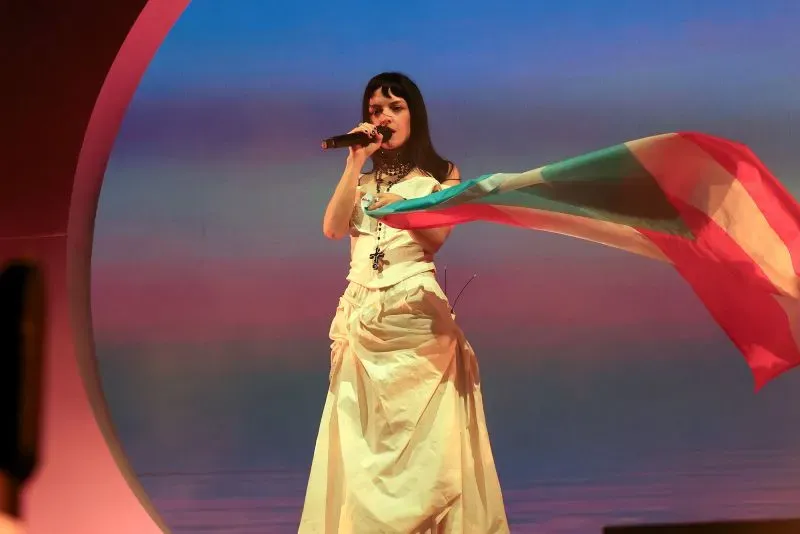 مغنية ترتدي فستانًا أبيض وتؤدي على المسرح، ممسكةً بعلم ملون، مع خلفية غامقة تعكس أجواء مهرجان كوتشيلا.