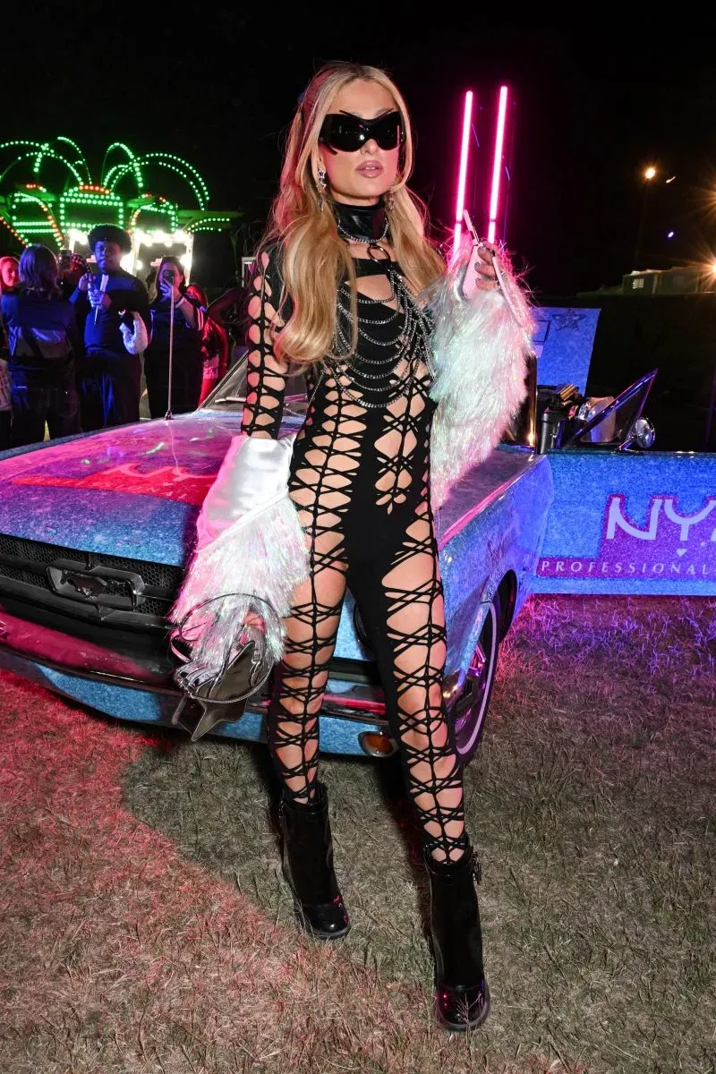 امرأة ترتدي فستانًا أسودًا مشدودًا مزينًا بأشرطة، مع نظارات شمسية كبيرة، وتقف بجانب سيارة زرقاء في مهرجان كوتشيلا.