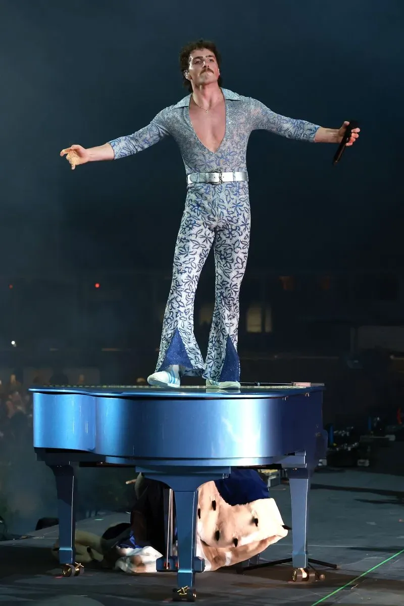 مغني يقف على بيانو أزرق، يرتدي زيًا لامعًا مزخرفًا، ويؤدي عرضًا حيويًا وسط أضواء المهرجان.