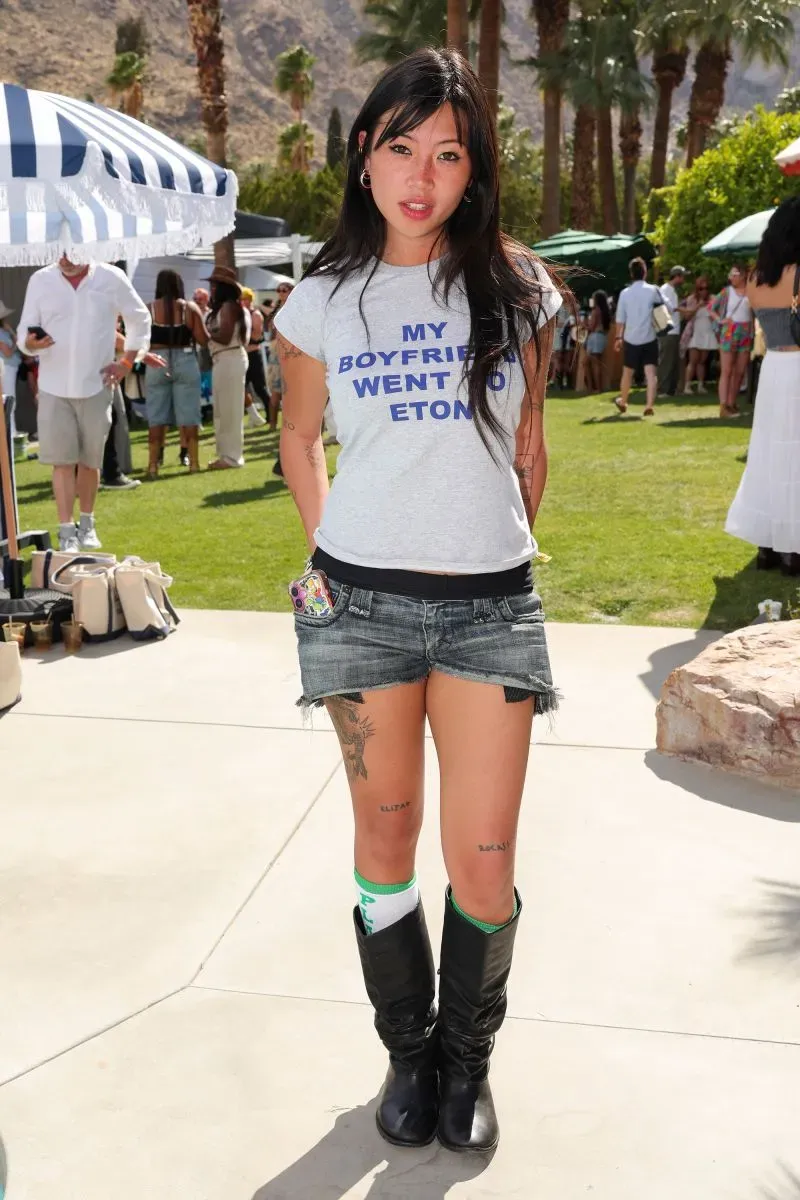 امرأة ترتدي قميصًا مكتوبًا عليه "MY BOYFRIEND WENT TO ETON" مع شورت جينز وقبعات طويلة، تتواجد في أجواء مهرجان كوتشيلا.