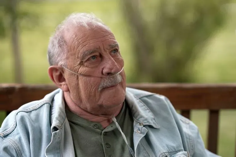 رجل مسن يرتدي قميصًا أزرقًا، يستخدم أنبوب أكسجين، يتأمل بقلق في الهواء الطلق، معبراً عن معاناته من مرض الرئة السوداء جراء عمله في تعدين الفحم.