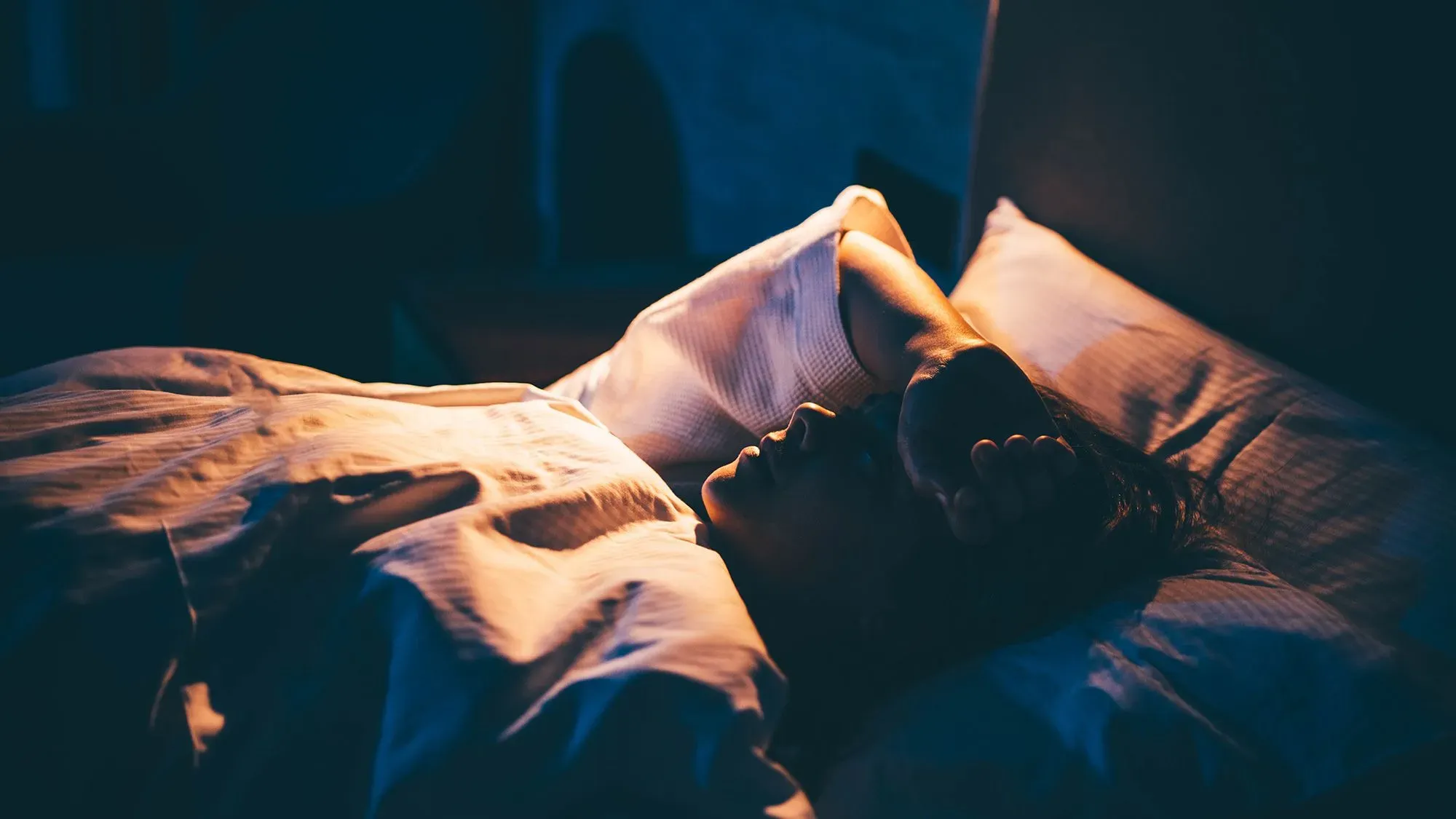 شخص مستلقٍ على السرير في غرفة مظلمة، يبدو عليه القلق وصعوبة النوم، مما يعكس موضوع الأرق والتقنيات لتحسين النوم.