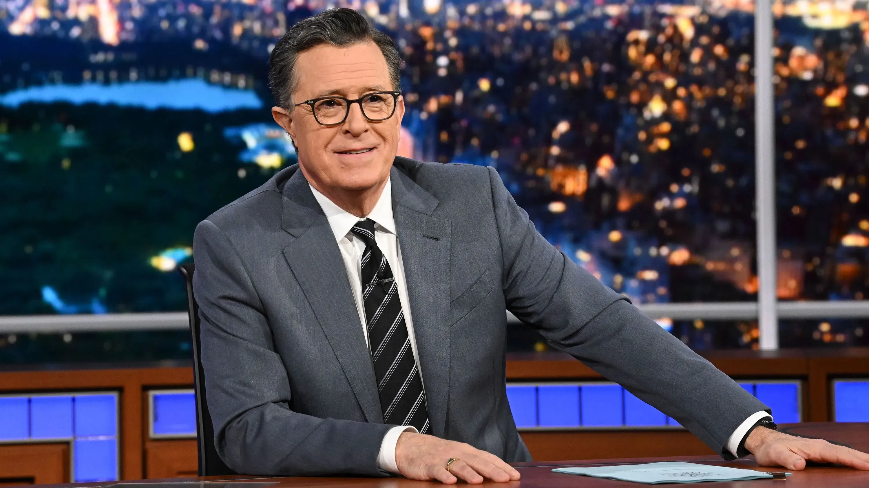 ستيفن كولبير يجلس على منصة برنامجه "العرض المتأخر" مع خلفية لمدينة مضاءة، استعدادًا لاستضافة ضيوفه في حلقة جديدة.