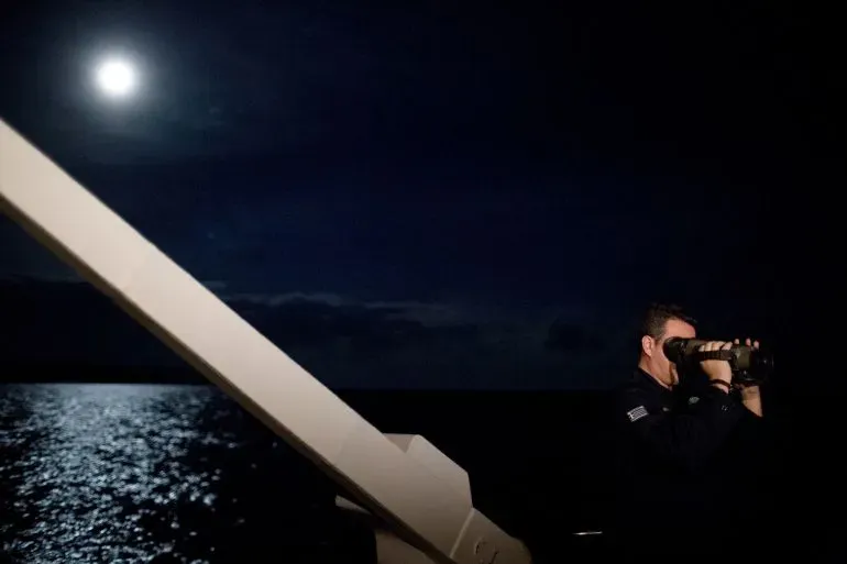 رجل من خفر السواحل يستخدم منظارًا لمراقبة البحر ليلاً، مع انعكاسات ضوء القمر على الماء، في سياق جهود البحث عن المهاجرين.